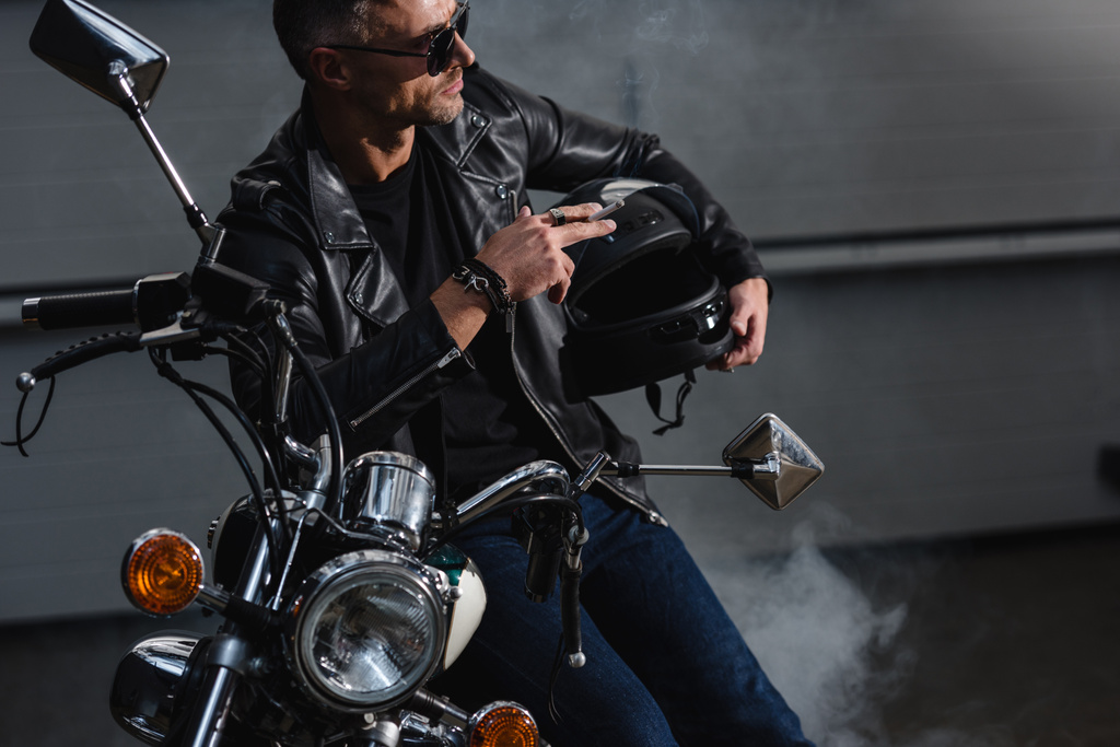 handsome motorbiker in sunglasses standing by motorcycle in garage and smoking cigarette - Photo, Image
