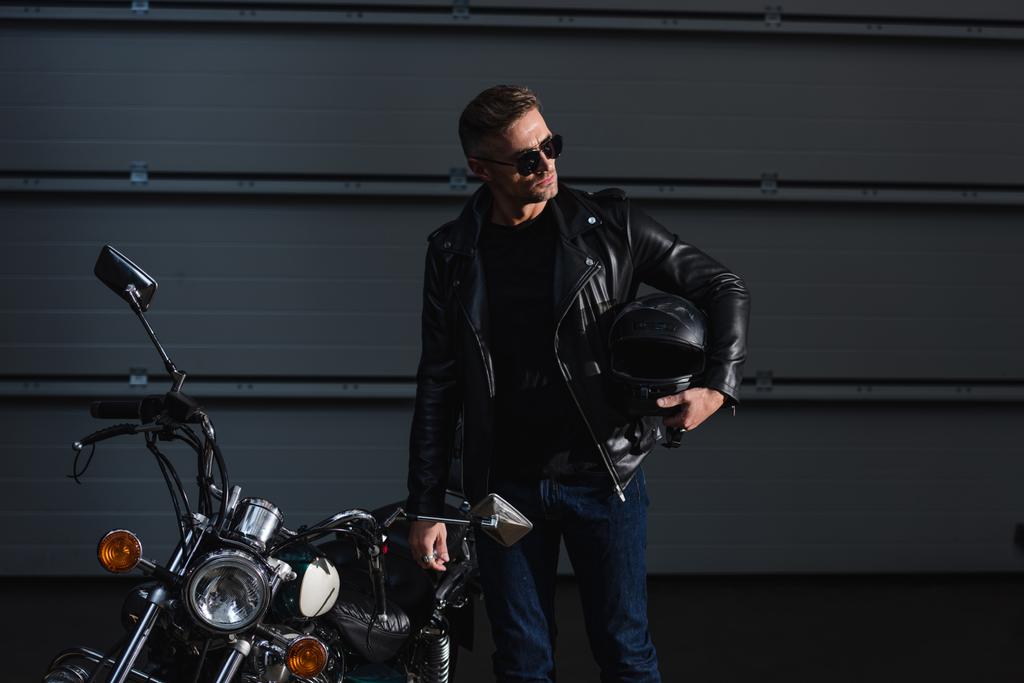 motero guapo en gafas de sol sosteniendo helment y de pie por motocicleta en garaje
 - Foto, imagen