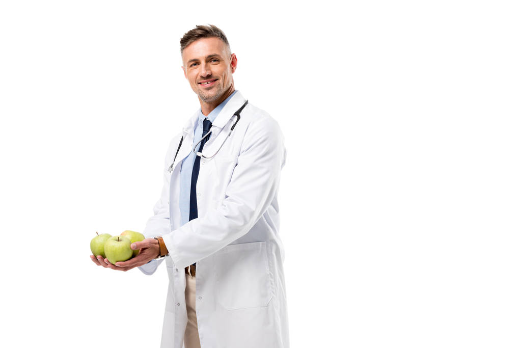 usmívající se lékař při pohledu na fotoaparát a drží čerstvá jablka izolovaných na bílém, zdravé stravovací koncept - Fotografie, Obrázek