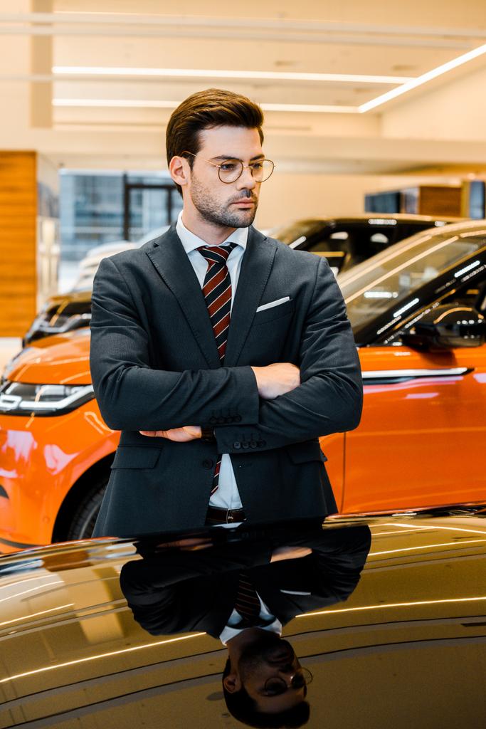 serio hombre de negocios en gafas posando con los brazos cruzados cerca de automóvil negro
 - Foto, Imagen