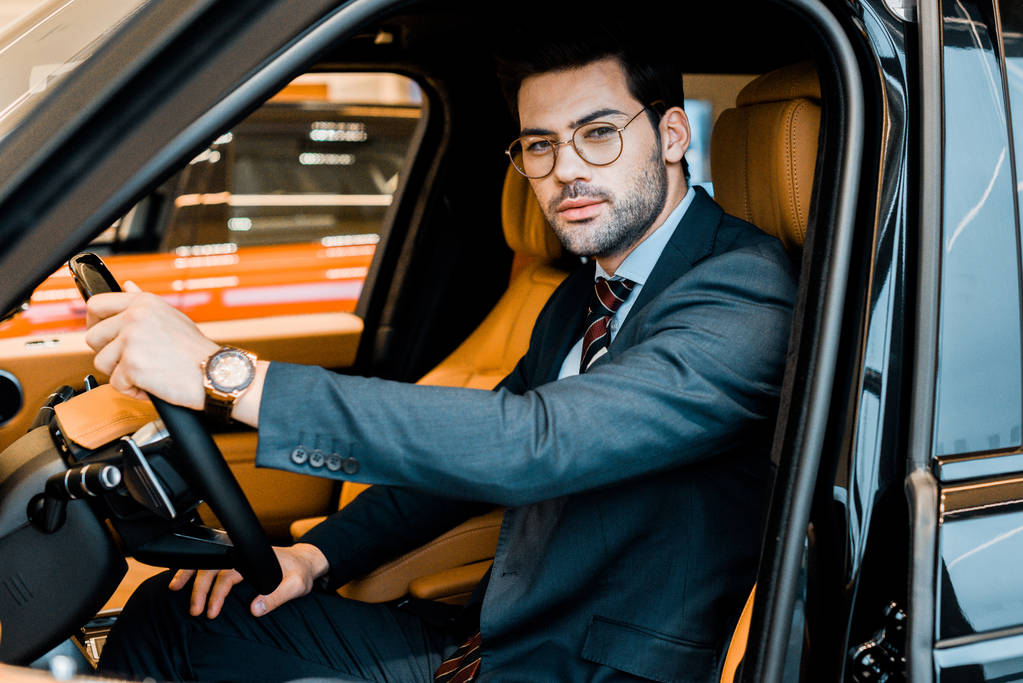 hombre de negocios con estilo en gafas de vista mirando a la cámara mientras está sentado en un coche de lujo
 - Foto, imagen