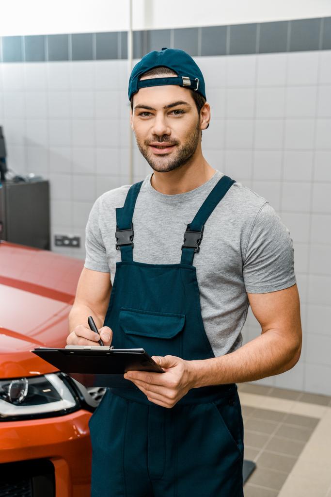 Porträt eines Automechanikers mit Notizblock, der in der Autowerkstatt in die Kamera schaut - Foto, Bild