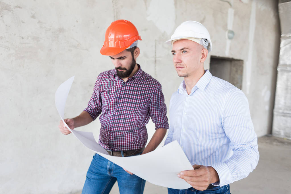 mannelijke ingenieurs, architecten in helmen staan bij het raam en houd een papierrol met een gebouw tekenen - Foto, afbeelding