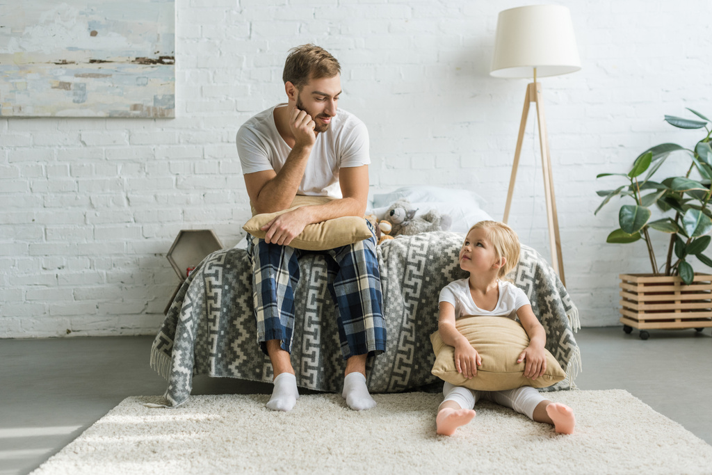father in pajamas sitting on bed and looking at cute little daughter sitting with pillow on carpet  - Photo, Image