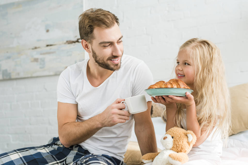 feliz padre en pijama sosteniendo la taza de té y mirando a la pequeña hija con sabrosos croissants en el plato
   - Foto, imagen