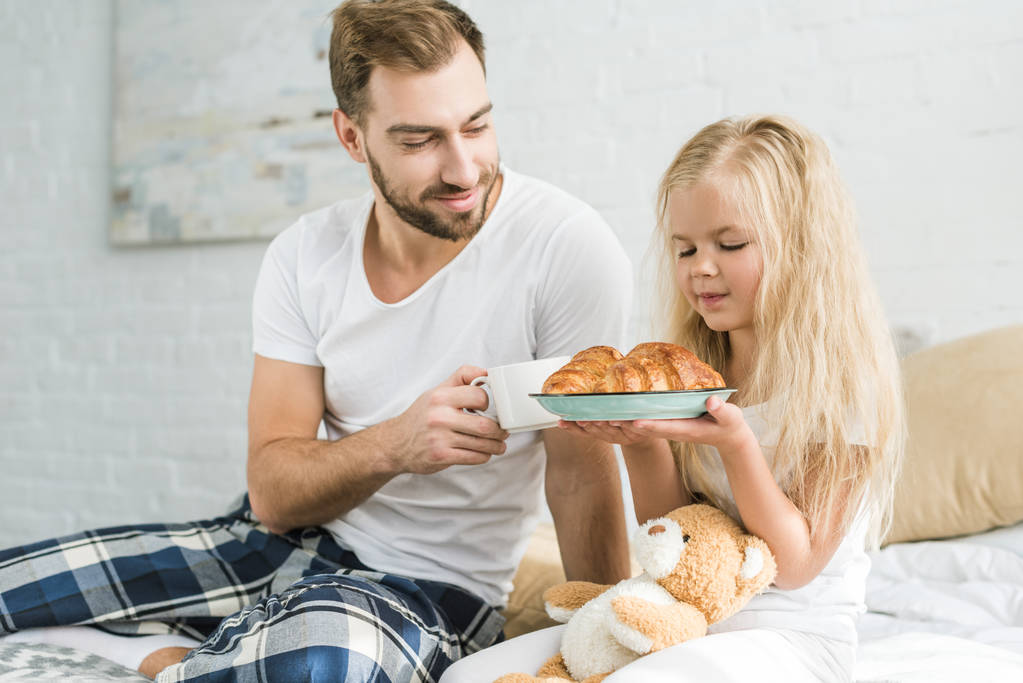 счастливый отец держит чашку кофе и смотрит на маленькую дочь с вкусными круассанами на тарелке
   - Фото, изображение