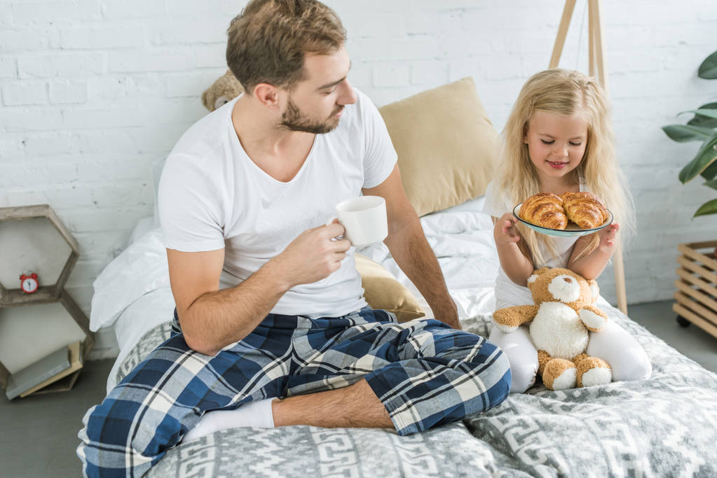 padre sosteniendo taza de café y mirando a la pequeña hija con sabrosos croissants en el plato
  - Foto, Imagen
