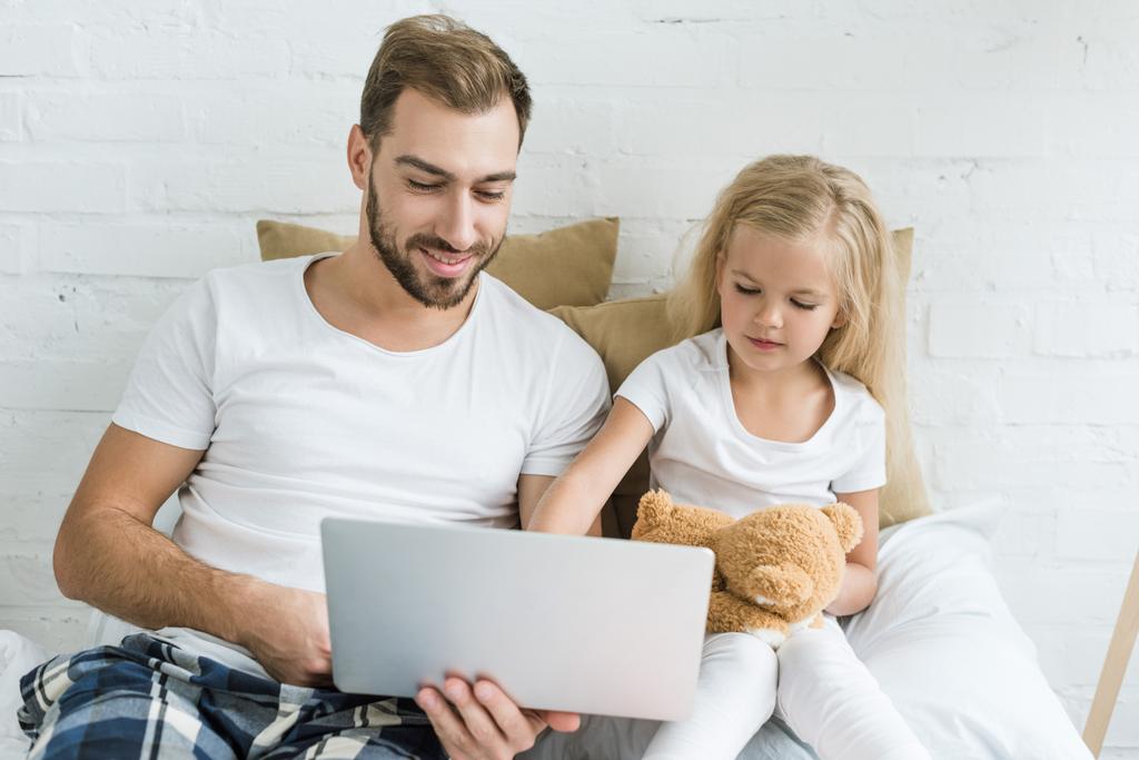 Šťastný otec a roztomilá malá dcera s teddy bear, společné používání notebooku v ložnici   - Fotografie, Obrázek