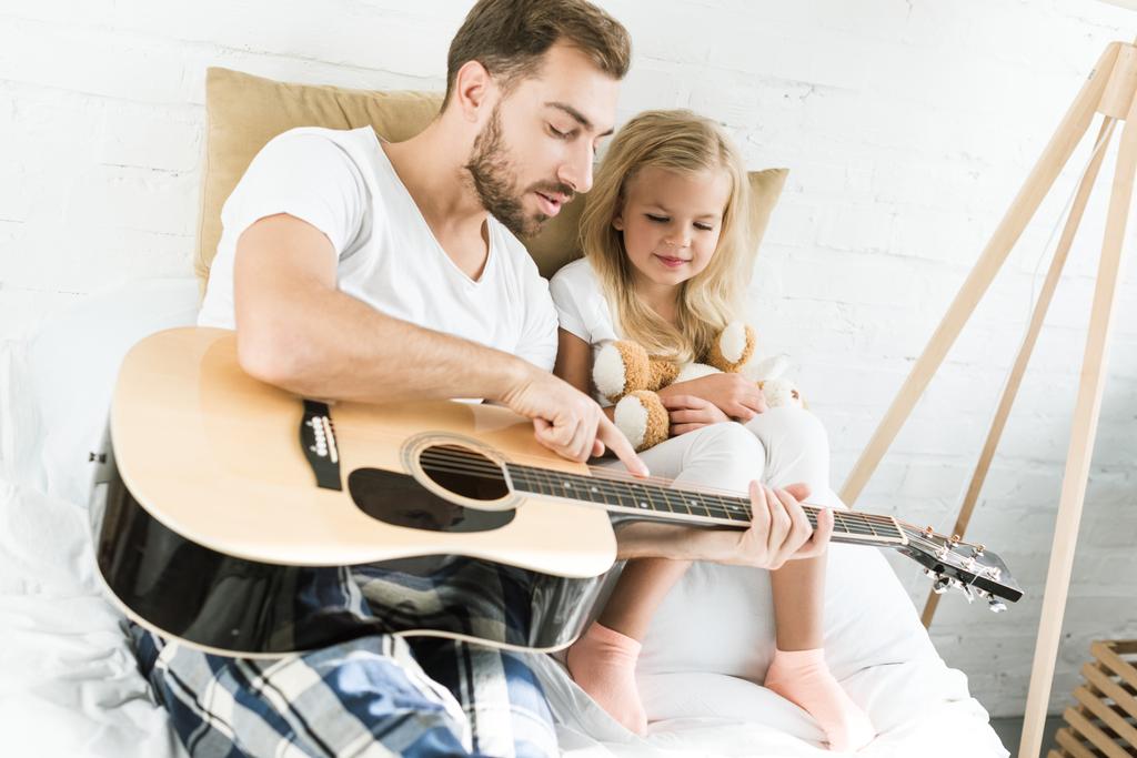père pointant vers la guitare acoustique à la petite fille souriante avec ours en peluche sur le lit
 - Photo, image