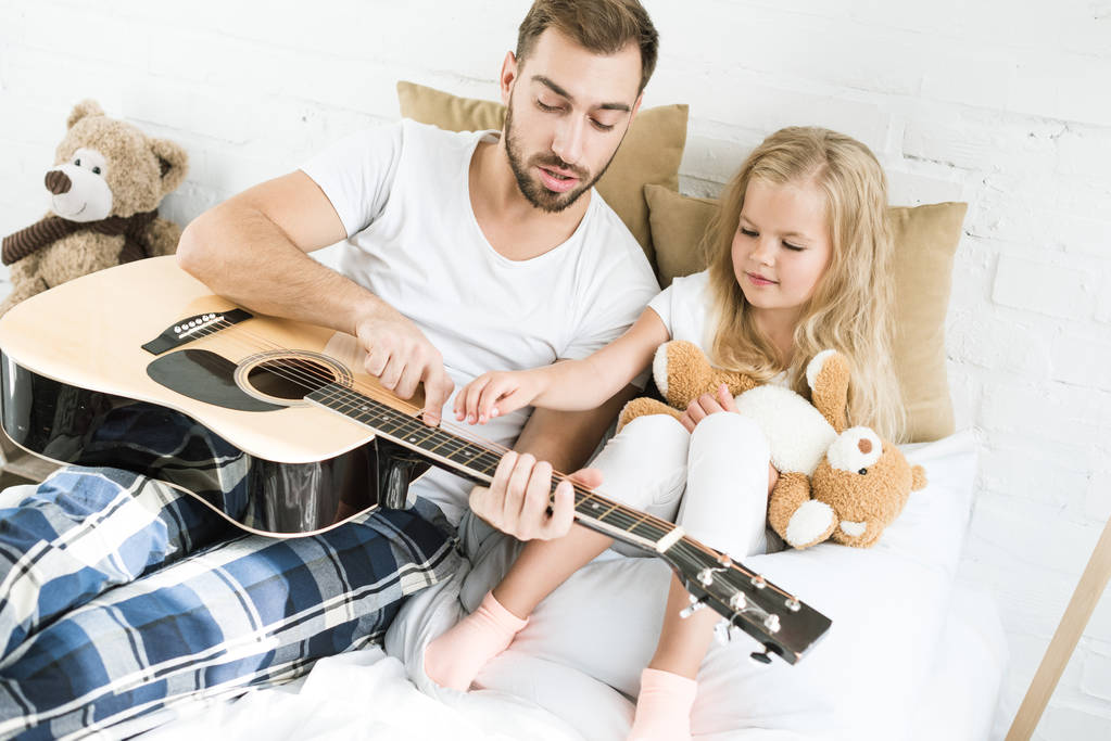 Blick aus der Vogelperspektive auf Vater und süße kleine Tochter mit Teddybär auf Akustikgitarre im Bett  - Foto, Bild