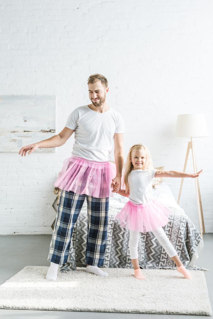 Gelukkig vader en schattig dochtertje in roze tutu rokken hand in hand en dansen samen thuis   - Foto, afbeelding