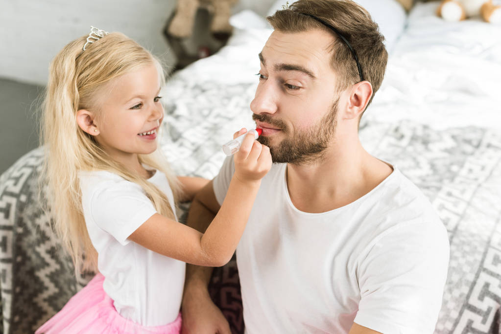 Entzückendes Töchterchen bringt zu Hause glücklichen bärtigen Vater mit rotem Lippenstift an     - Foto, Bild