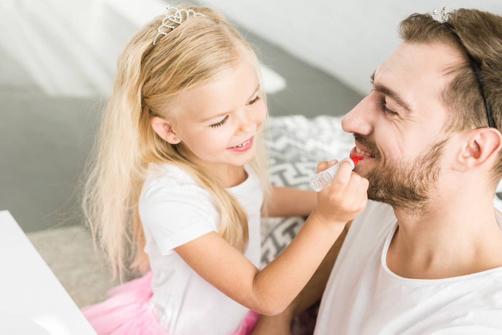 adorable little daughter applying makeup to happy father at home  - Photo, Image