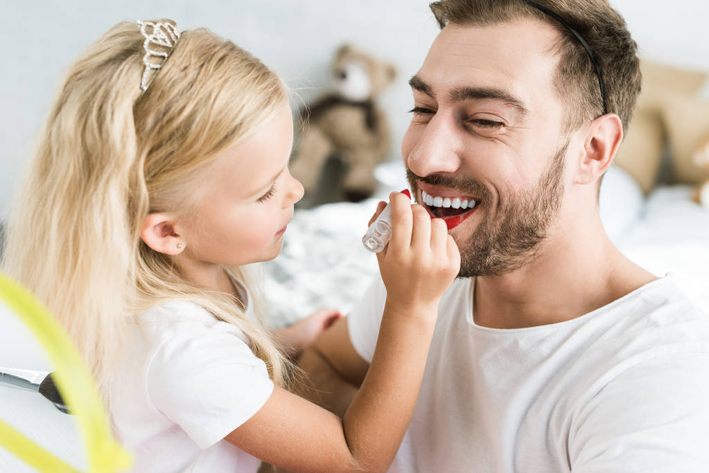 schattig dochtertje rode lippenstift op gelukkig bebaarde vader toe te passen - Foto, afbeelding