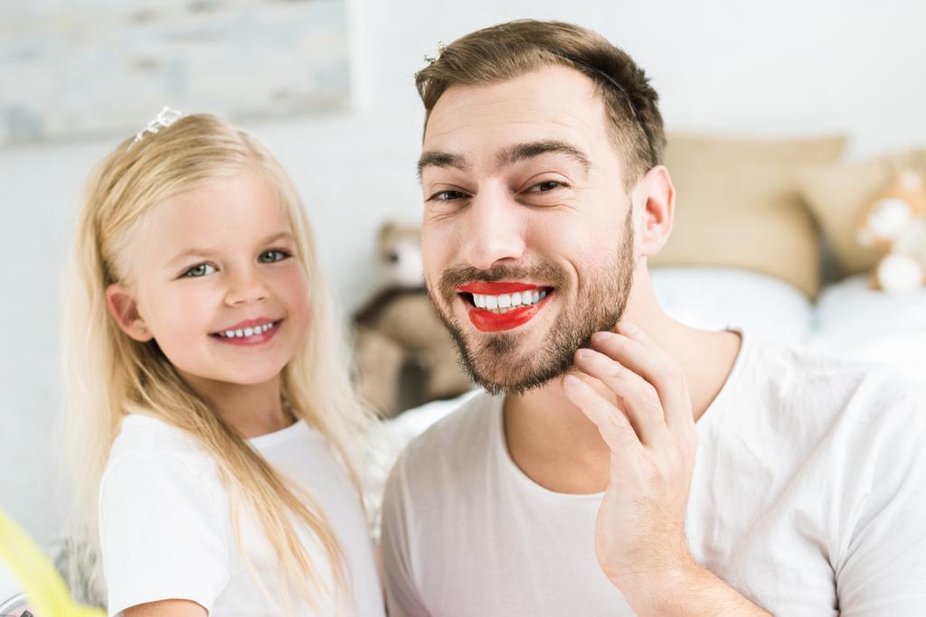 Entzückende kleine Tochter und glücklicher bärtiger Vater mit rotem Lippenstift, der in die Kamera lächelt  - Foto, Bild