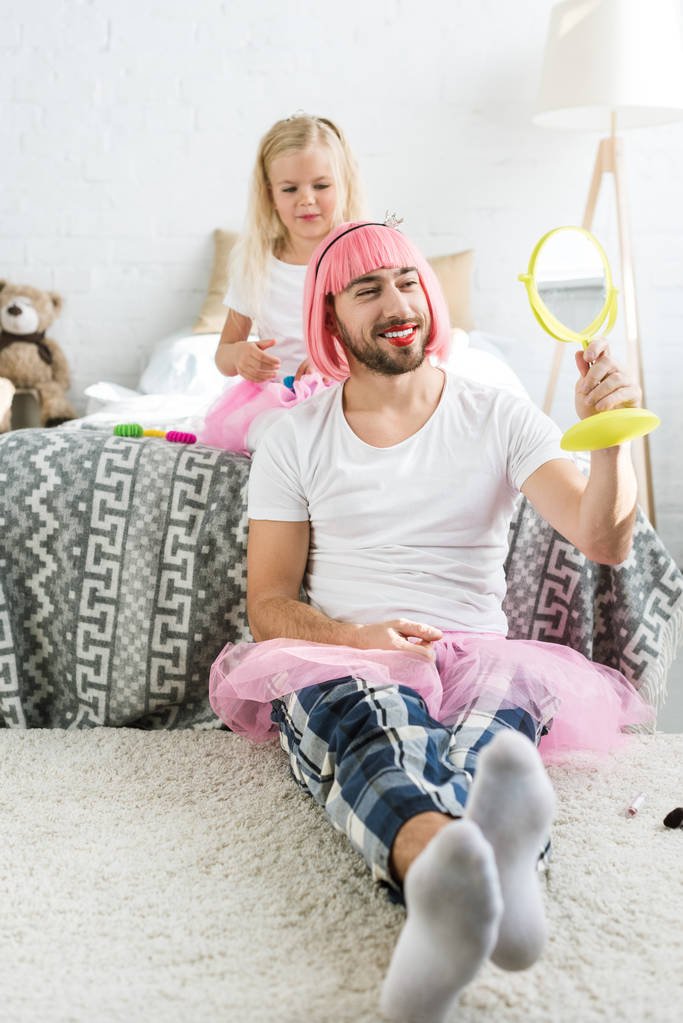 entzückende kleine Tochter spielt mit glücklichem Vater in pinkfarbener Perücke und Tutu-Rock und schaut in den Spiegel      - Foto, Bild
