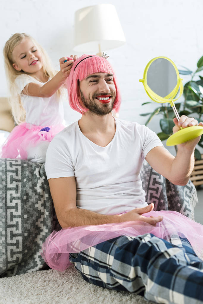 söpö pieni tytär tutu hame leikkii hymyilevä isä vaaleanpunainen peruukki katselee peiliin
    - Valokuva, kuva