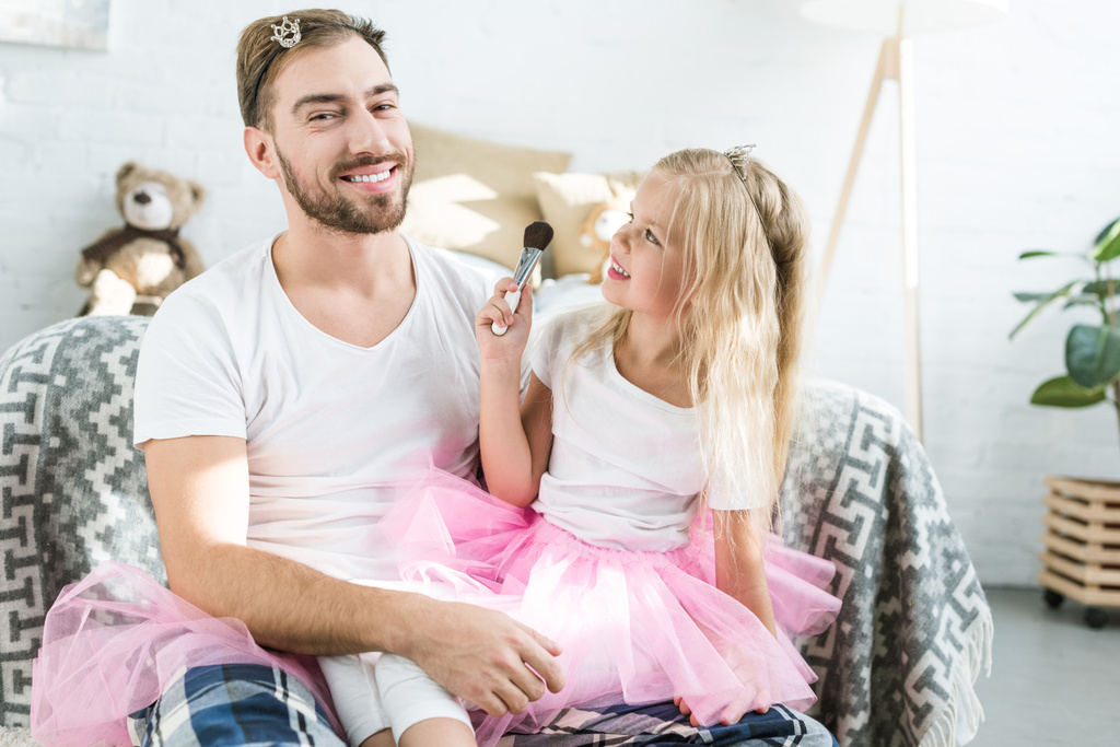adorable little daughter in pink tutu skirt applying makeup to happy father  - Photo, Image