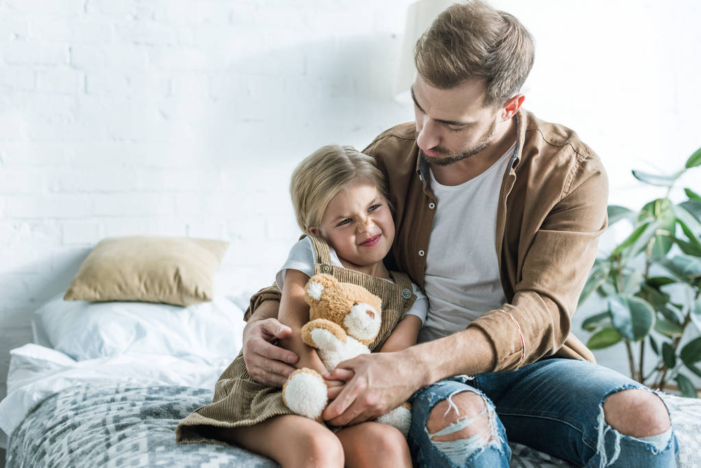 Baba küçük kızı yatakta oyuncak ayı ile oturan bakarak  - Fotoğraf, Görsel