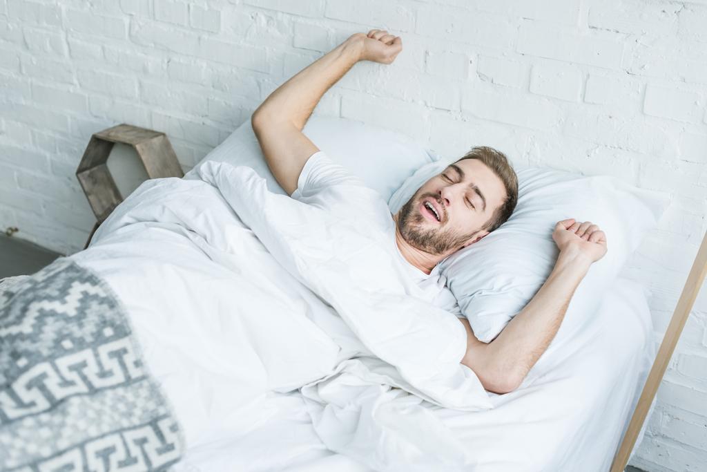korkea kulma näkymä nuori mies haukottelu ja venyttely sängyssä
  - Valokuva, kuva