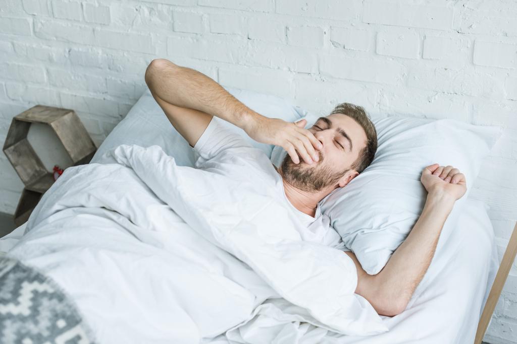 όμορφος νεαρός άνδρας χασμουρητό και το τέντωμα ξαπλωμένοι στο κρεβάτι  - Φωτογραφία, εικόνα