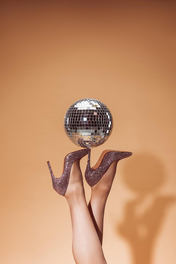 bijgesneden afbeelding van vrouw met glanzende disco bal op hoge hakken aan partij op beige - Foto, afbeelding