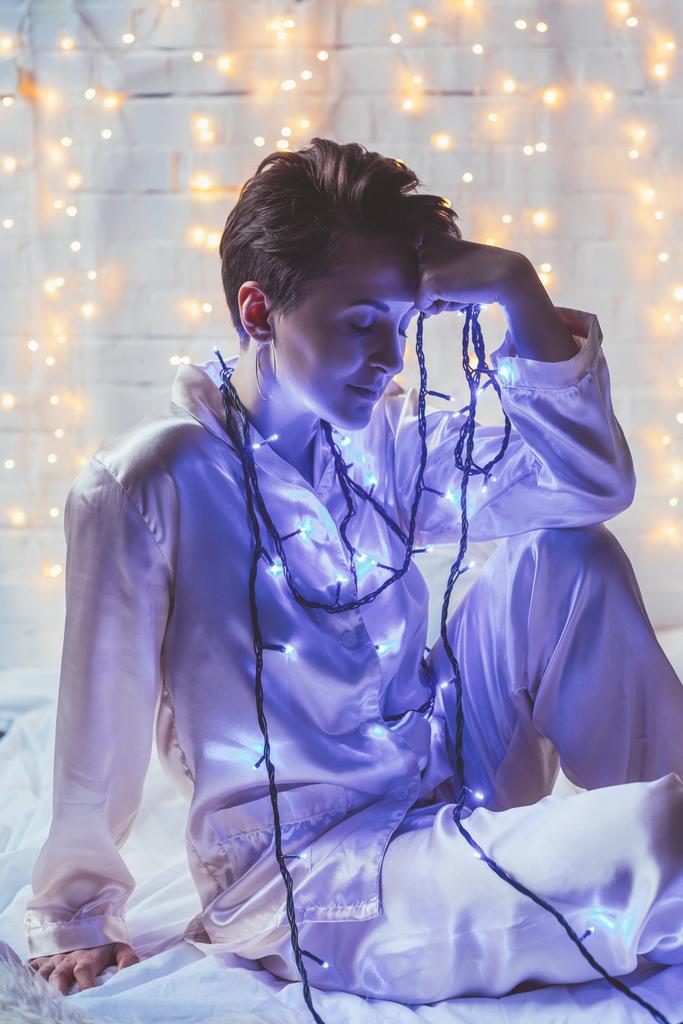 задумчивая женщина в пижаме с рождественскими огнями отдыхает на кровати дома
 - Фото, изображение