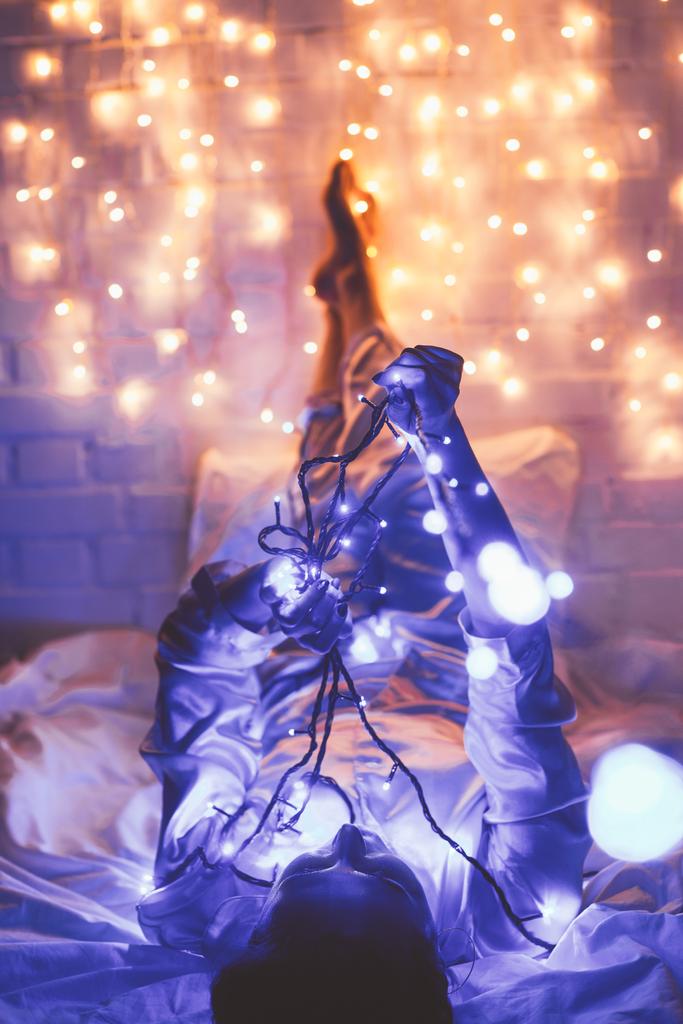 женщина в пижаме отдыхает на кровати с рождественскими огнями вокруг
 - Фото, изображение