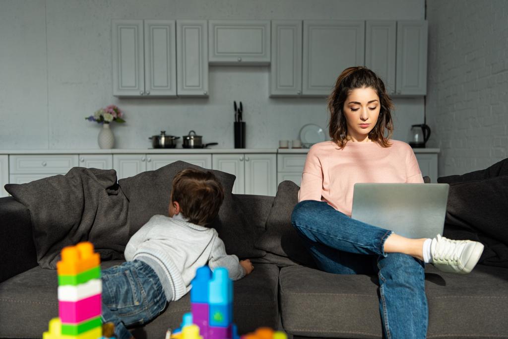 rear view of little boy climbing on sofa while his mother using laptop at home - Photo, Image