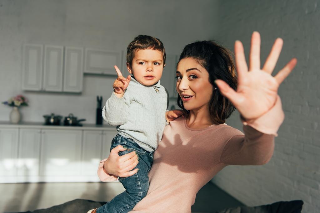 heureuse femme attrayante tenant petit fils et agitant à la main à la maison
 - Photo, image
