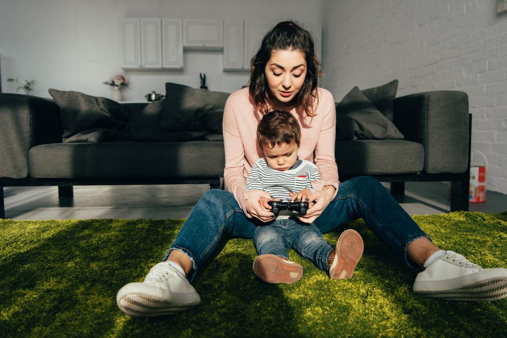 cute child and his mother sitting on floor and playing video game with joystick at home - Photo, Image