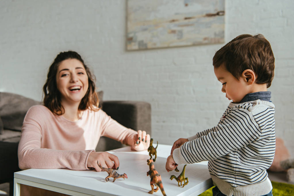 Lachende Frau und kleiner Sohn spielen Spielzeug-Dinosaurier am Tisch im heimischen Wohnzimmer - Foto, Bild