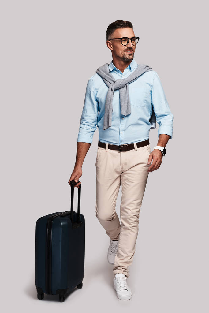 voller Länge gut aussehender junger Mann, der vor grauem Hintergrund mit Reisetasche läuft  - Foto, Bild