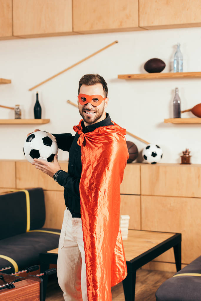 giovane uomo sorridente in costume da supereroe rosso con pallone da calcio nel caffè
 - Foto, immagini
