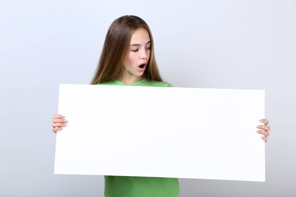 Jovencita sorprendida sosteniendo tablero en blanco sobre fondo gris
 - Foto, Imagen