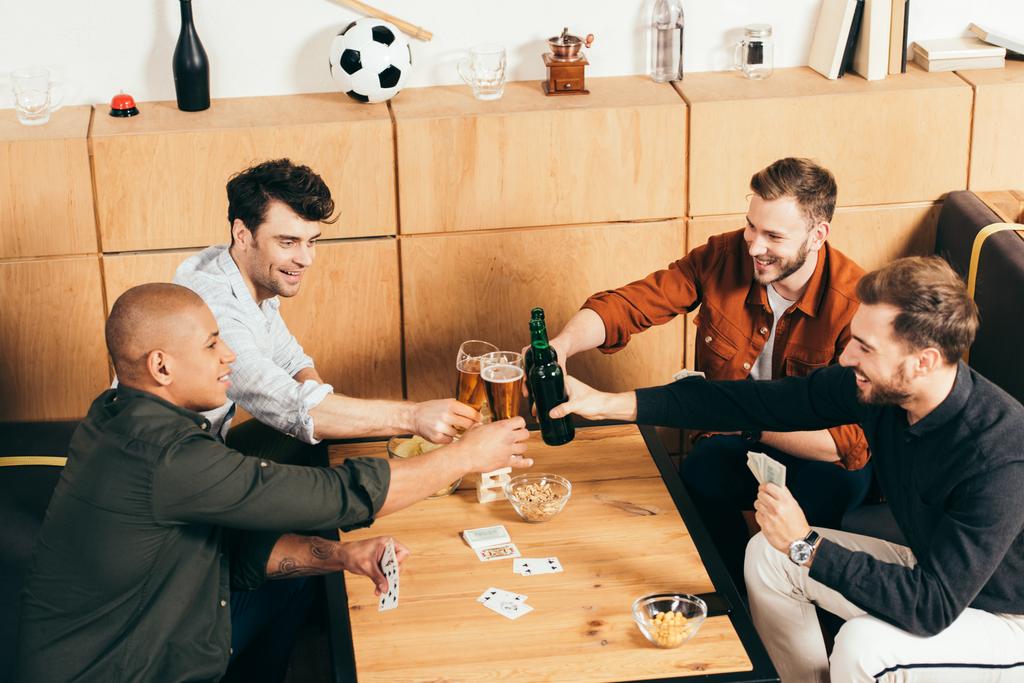 smiling multiracial friends clinking drinks at tabletop with cards and snacks in cafe - Photo, Image