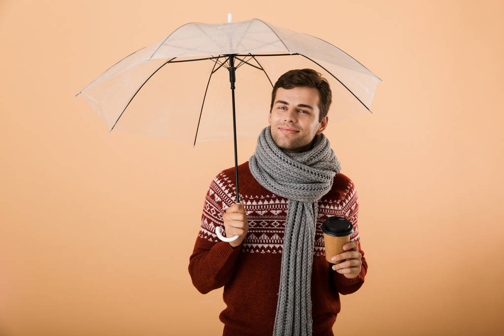 Retrato de um jovem feliz vestido com suéter e cachecol isolado sobre fundo bege, de pé sob guarda-chuva, segurando xícara de café
 - Foto, Imagem