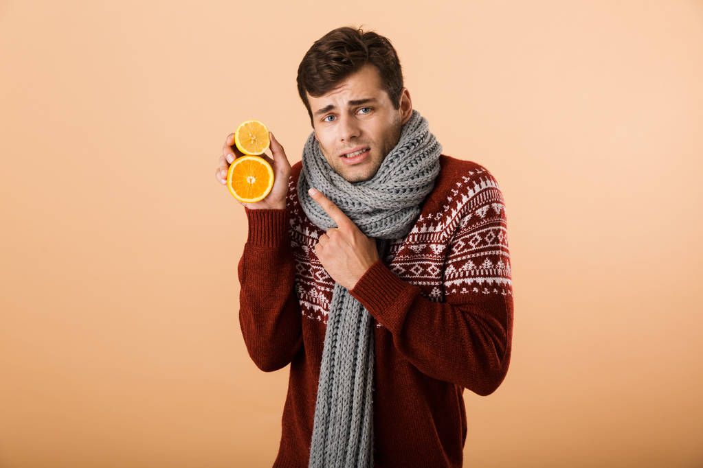 Portrait a sick man dressed in sweater and scarf isolated over beige background, holding sliced oranges - Photo, Image