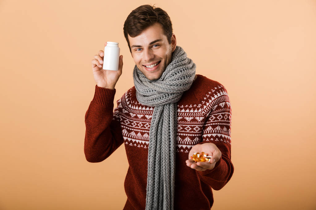 Porträt eines lächelnden Mannes in Pullover und Schal vor beigem Hintergrund, der Flasche und Pillen in der Hand hält - Foto, Bild