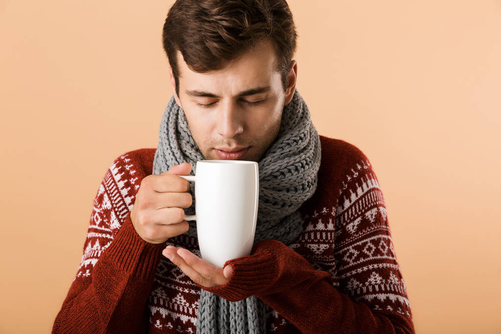 Close up of a handsome young man dressed in sweater and scarf drinking tea from a cup isolated over beige background - Photo, Image