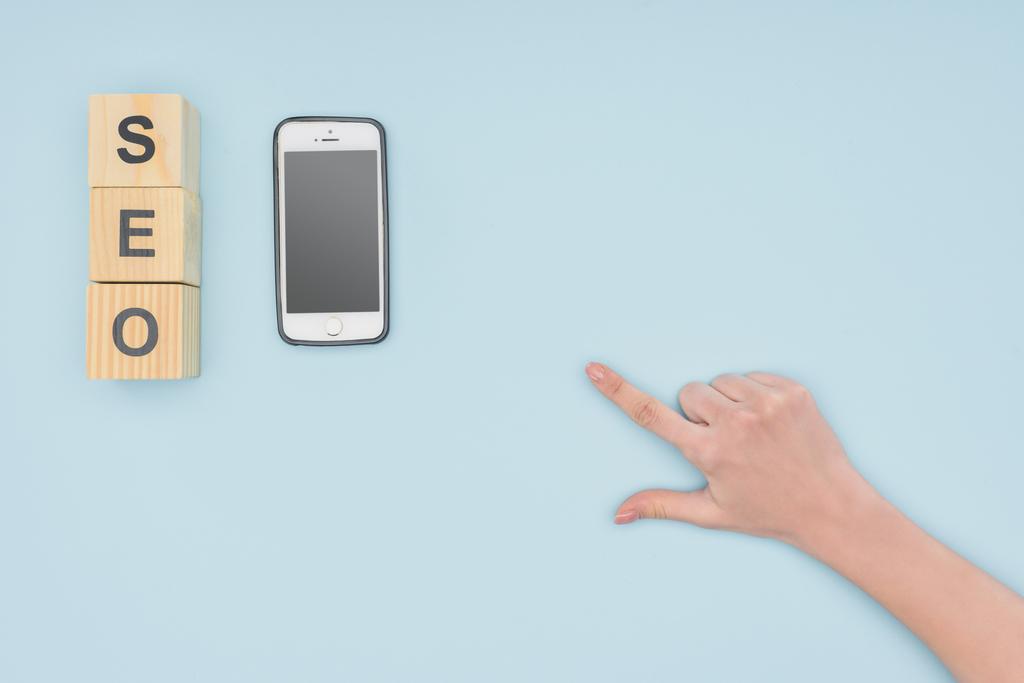 Top view of female hand pointing at smartphone and wooden cubes on light blue background  - Photo, Image