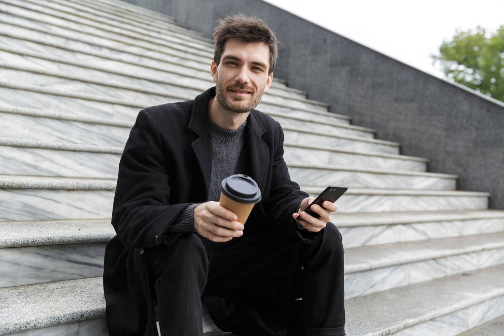 Wizerunek przystojny młody biznes człowiek siedzi na zewnątrz za pomocą telefonu komórkowego, picia kawy. - Zdjęcie, obraz