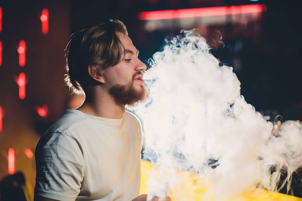 Young handsome hipster man with beard sitting in cafe with a cup of coffee, vaping and releases a cloud of vapor. - Photo, Image