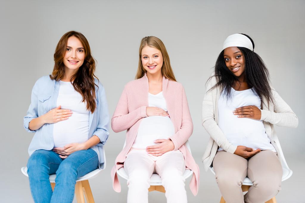 беременные многонациональные женщины, сидящие на стульях и трогательные животы, изолированные на сером
 - Фото, изображение
