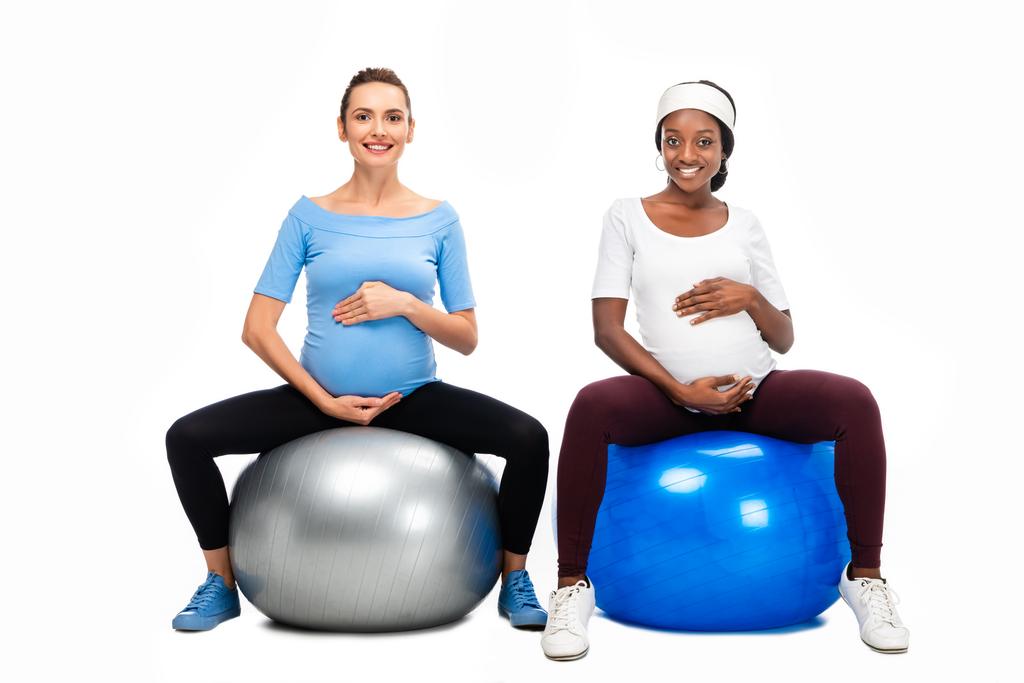twee multiculturele zwangere vrouwen zit op fitness ballen en hand in hand op buiken geïsoleerd op wit - Foto, afbeelding