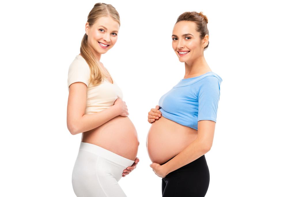 twee zwangere vriendinnen buiken in profiel en glimlachend geïsoleerd op wit tonen - Foto, afbeelding