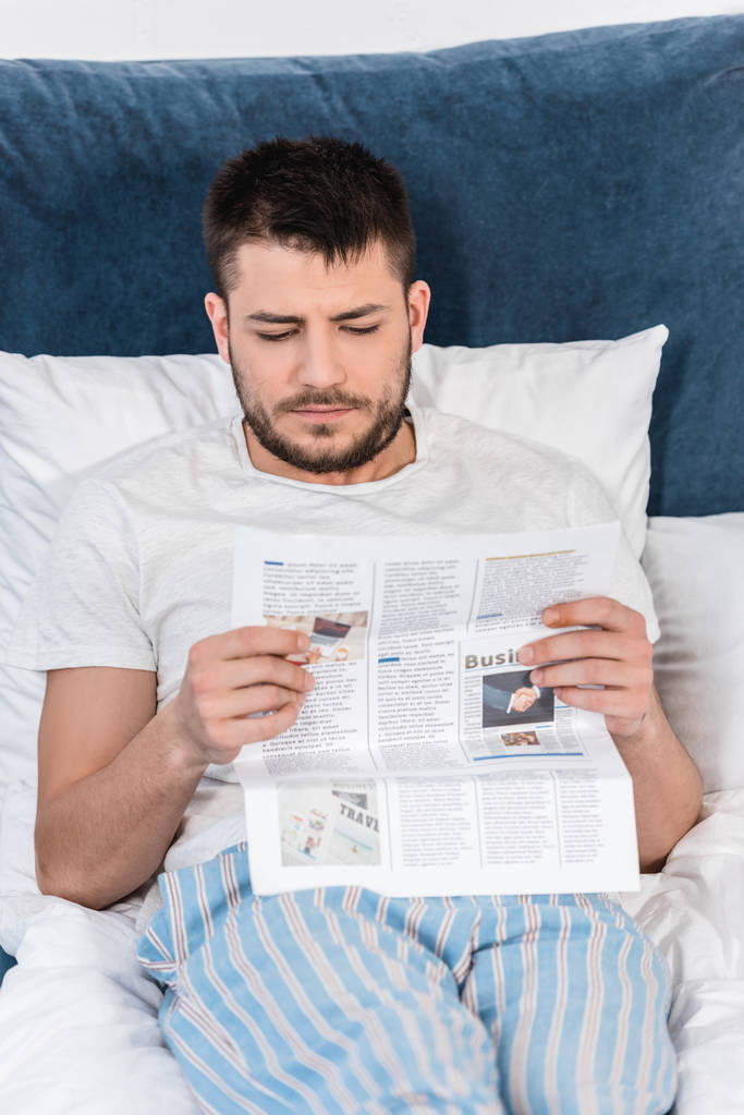przystojny mężczyzna w piżamie czytanie gazety w łóżku rano w domu - Zdjęcie, obraz
