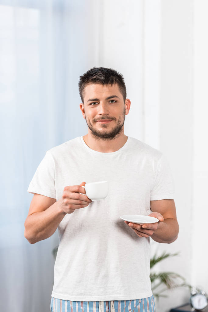 handsome man in shirt and pajamas holding cup of coffee and looking at camera in morning in bedroom - Photo, Image