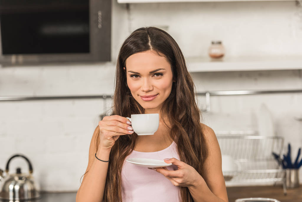 όμορφη γυναίκα χαρούμενη κρατώντας το φλιτζάνι του καφέ και κοιτάζοντας κάμερα πρωί στην κουζίνα - Φωτογραφία, εικόνα