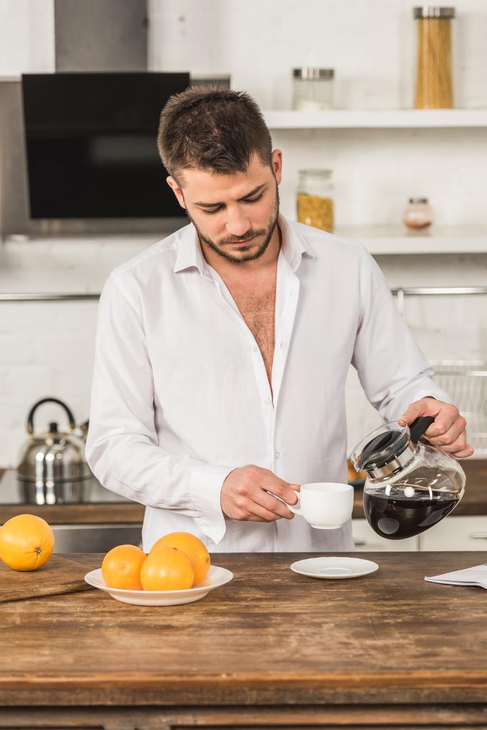 schöner Mann im Hemd, der morgens in der Küche Kaffee in Tasse einschenkt - Foto, Bild